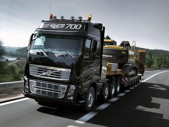 Motgående svart Volvo lastebil med gravemaskin på lasteplanet